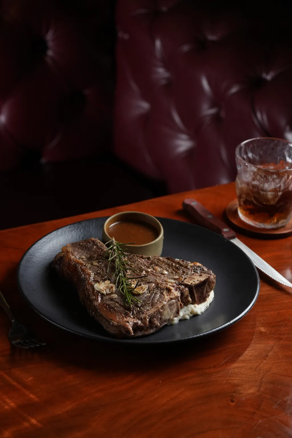 A Ninan's T-bone steak on a black plate with mash, rosemary and a pot of sauce